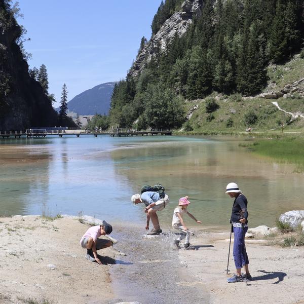 Four people are enjoying a sunny day by a clear, turquoise mountain lake, with two children and an adult splashing in the shallow water